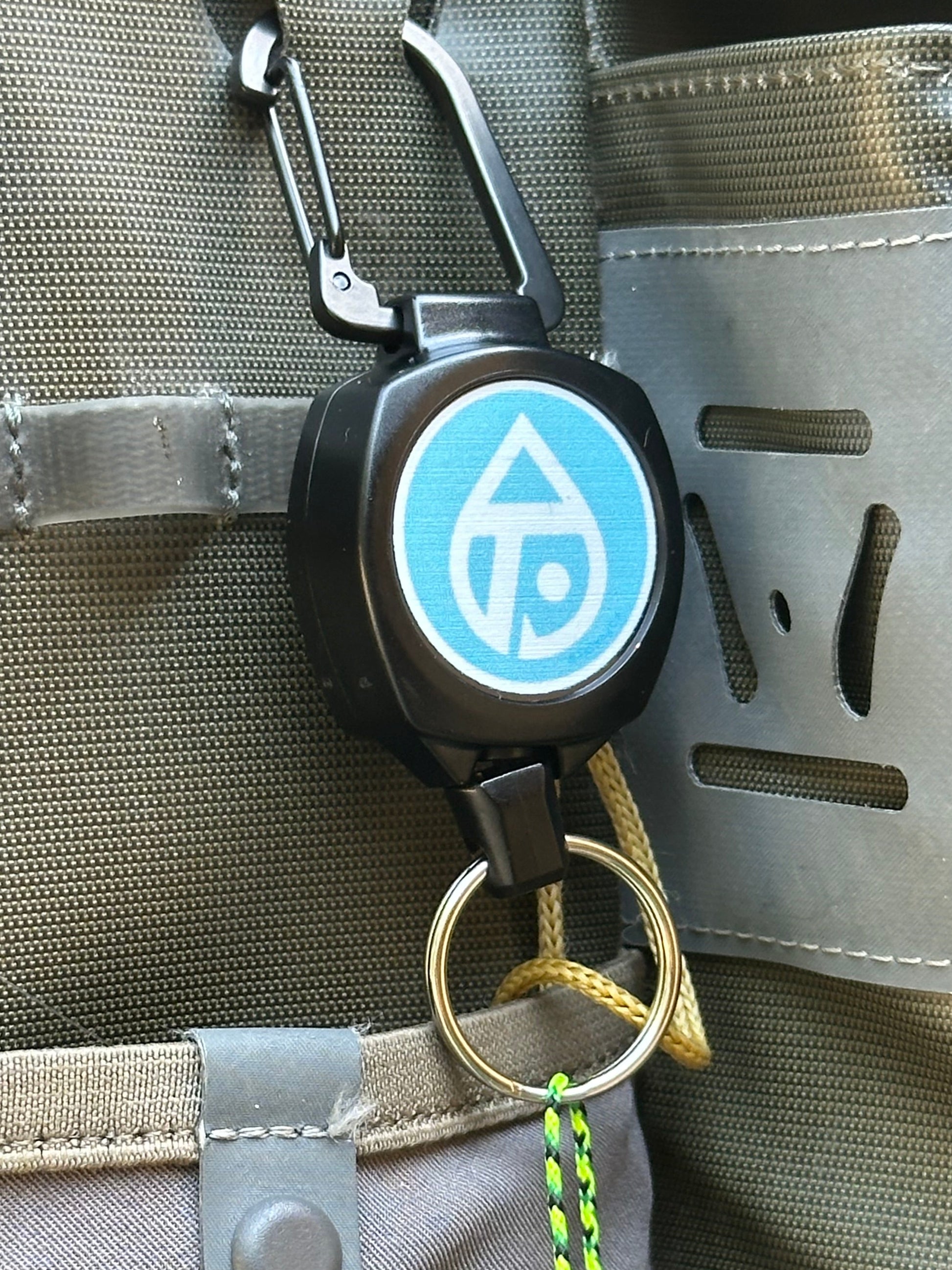 A close-up of a black retractable keychain bag with a blue and white logo, attached to a gray outdoor gear bag. A bright green and black braided lanyard hangs from the key ring.