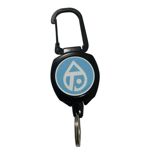A black retractable fishing zinger with a blue and white circular logo on the face, attached to a silver metal key ring, photographed on a plain light gray surface.
