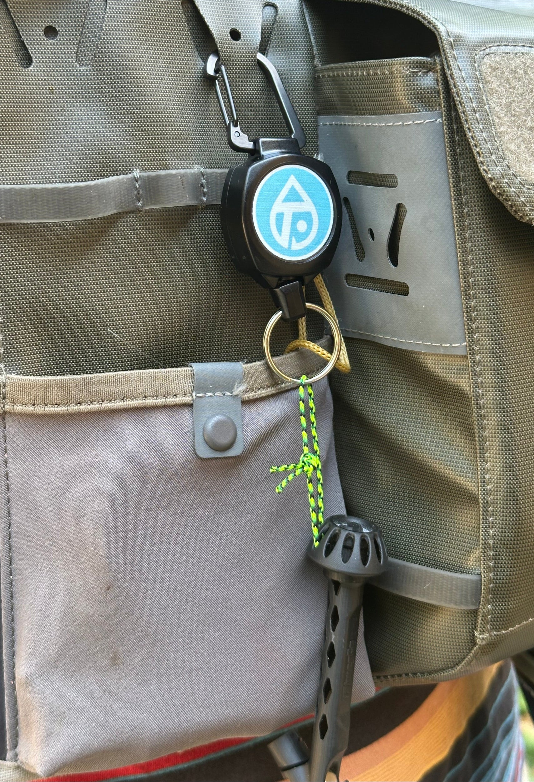 A close-up of a green and gray fishing tackle bag with a black keychain featuring a blue circular logo, attached to a carabiner on the bag's side.